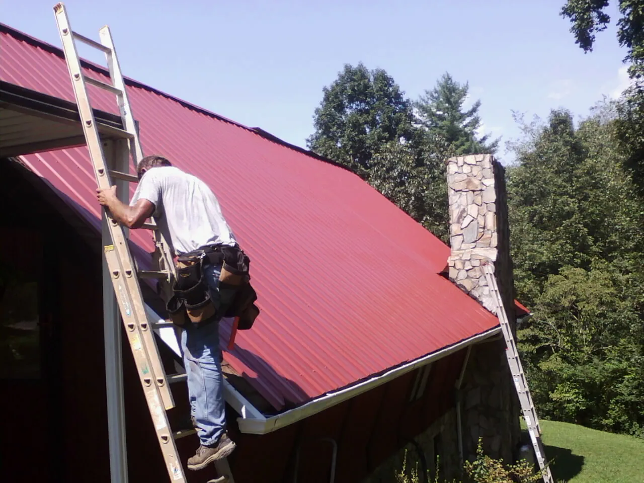 Roofer on a ladder beside a red metal roof and stone chimney — Soffit & Fascia Repair by Roofing Brothers in Blythewood